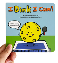 Load image into Gallery viewer, A hand holding a children's board book titled 'I Dink I Can!' against a plain white background. The book cover features a cartoon yellow dinking ball character standing on a paddle net with a bright blue sky and green ground background.
